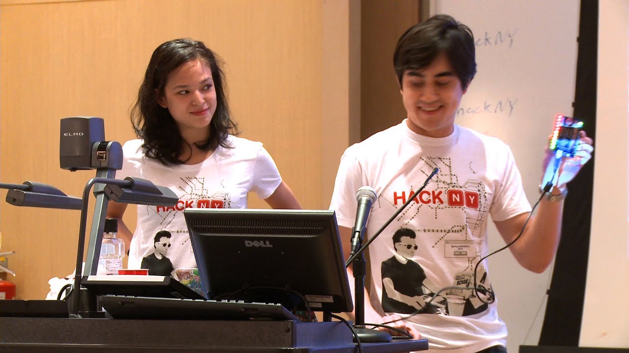 Two young people wearing HackNY t-shirts speak at a presentation podium with microphones during what appears to be the HackNY Spring 2013 event. A laptop and presentation materials are visible on the podium.