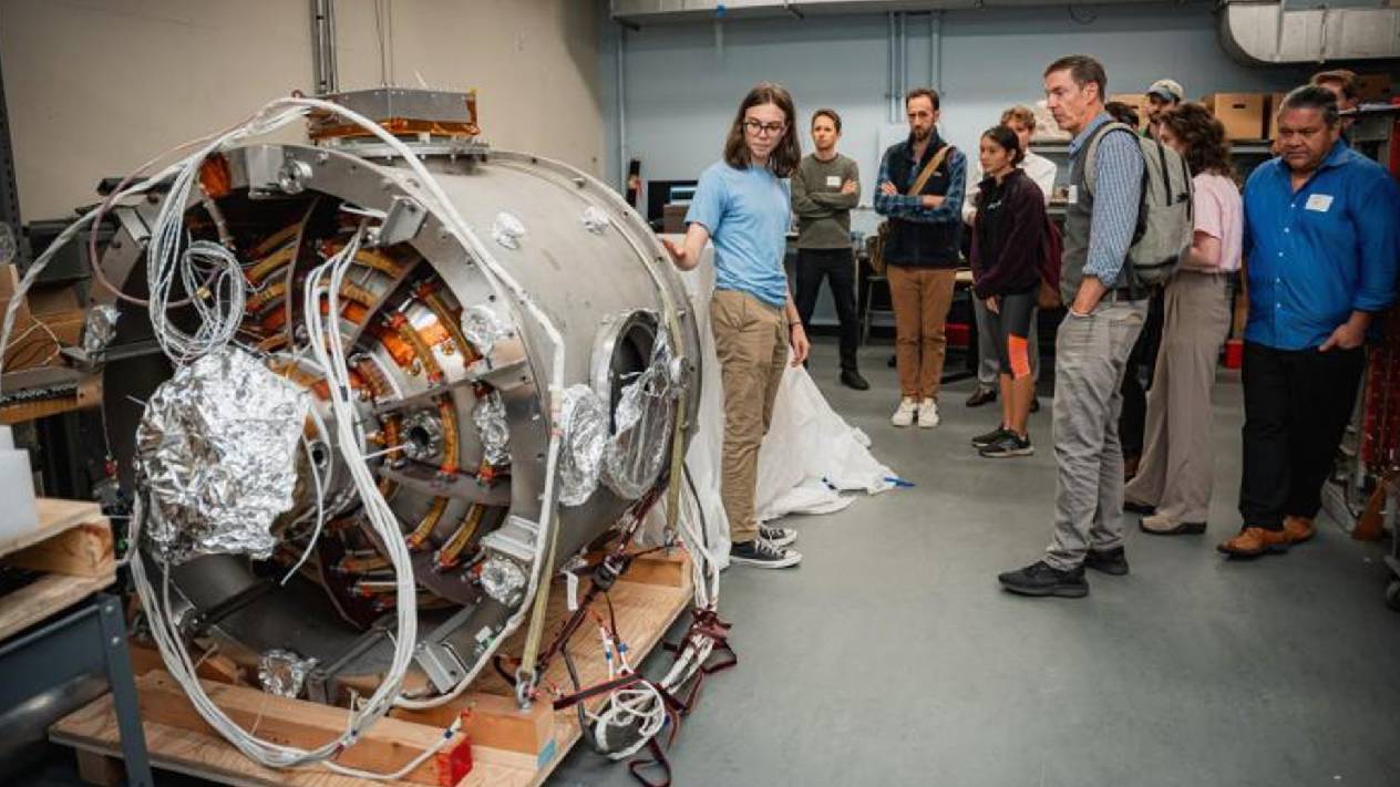 A group of people tours a laboratory, gathered around a large cylindrical scientific device with exposed coils, wiring, and reflective insulation. A woman in a blue shirt stands beside the equipment, explaining or demonstrating it to the group.