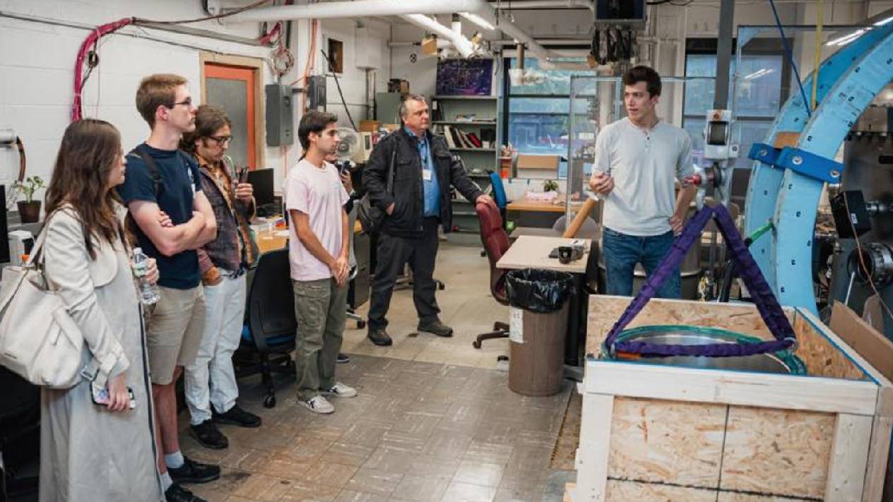 A group of people tours a laboratory, gathered around a large cylindrical scientific device with exposed coils, wiring, and reflective insulation. A man in a light blue shirt stands beside the equipment, explaining or demonstrating it to the group.