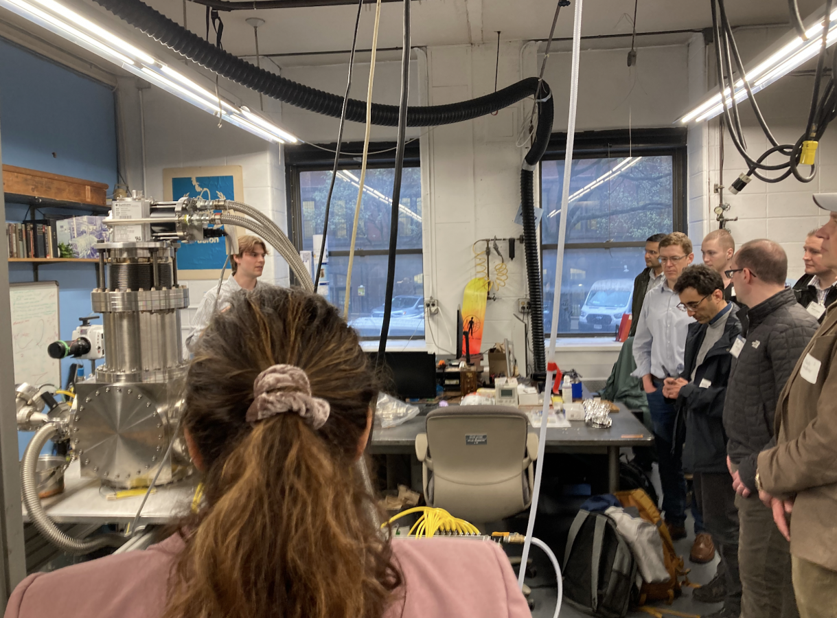 In a laboratory, a group of people stands around a workbench listening to someone explain equipment; a large stainless-steel vacuum chamber with hoses is on the left, and a person with a ponytail is in the foreground facing the group.