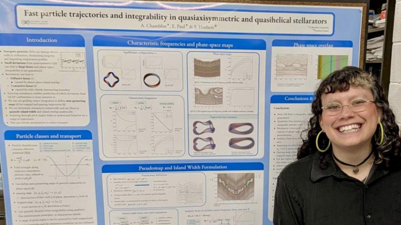 Smiling person with curly hair and glasses stands beside a large blue-and-white scientific poster filled with graphs and diagrams about particle trajectories and integrability.