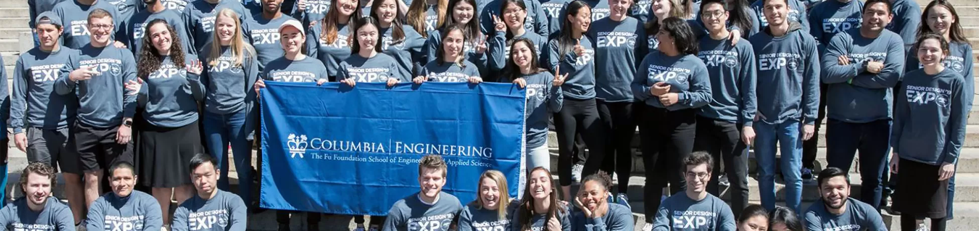 a large group of students wearing shirts that say 2019 senior design expo sit outside on the steps in front of Columbia's Low Library and a statue of alma mater
