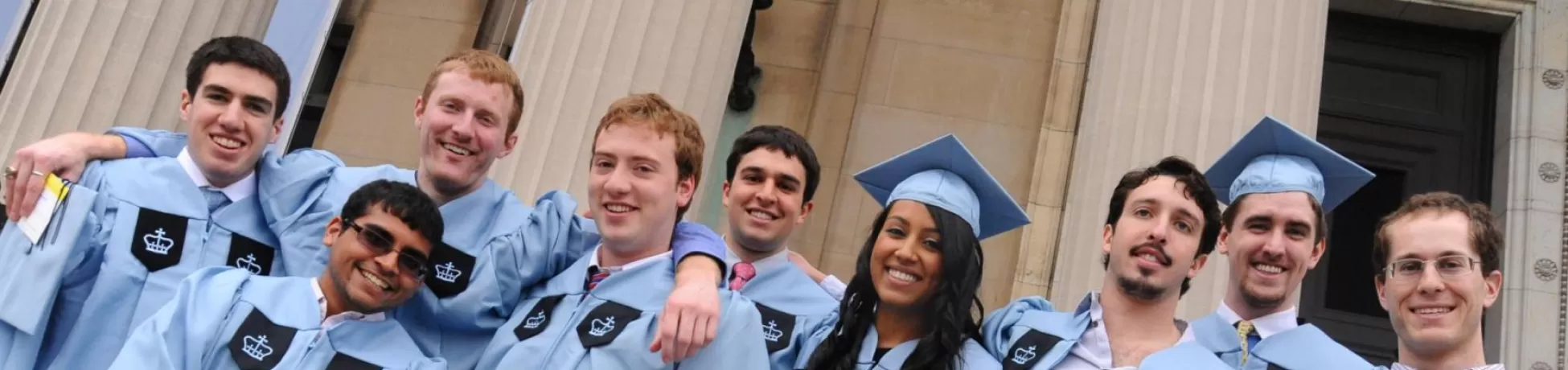 Group of smiling graduates in light blue caps and gowns standing together in front of a building with tall stone columns.