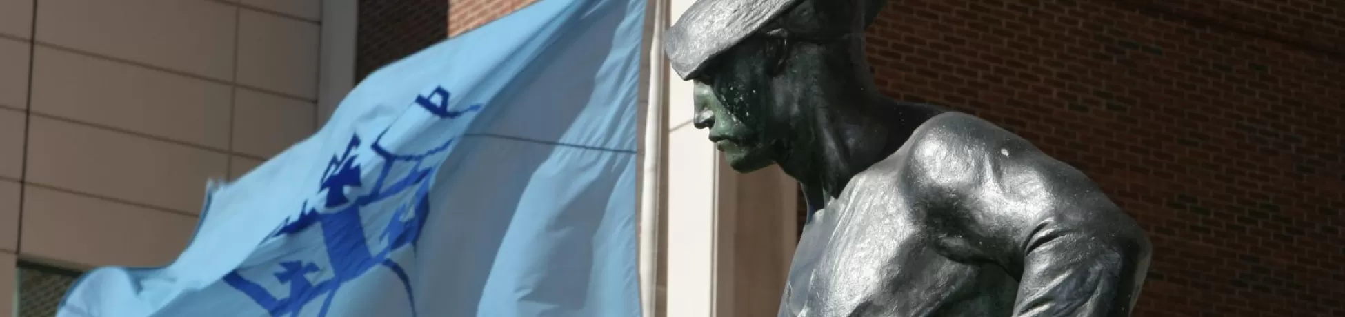 Bronze statue of a man wearing a cap, looking downward with hands on hips, in front of a brick building with a blue flag waving on a pole nearby.