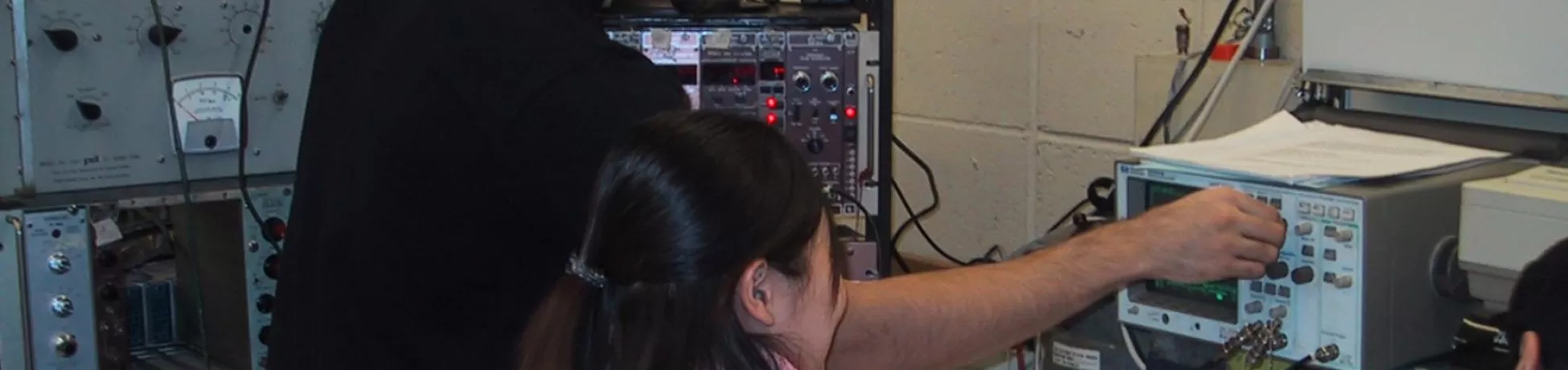 Two students in a lab adjust knobs on an oscilloscope and electronic testing equipment while reviewing notes at a cluttered workstation with cables and instruments.