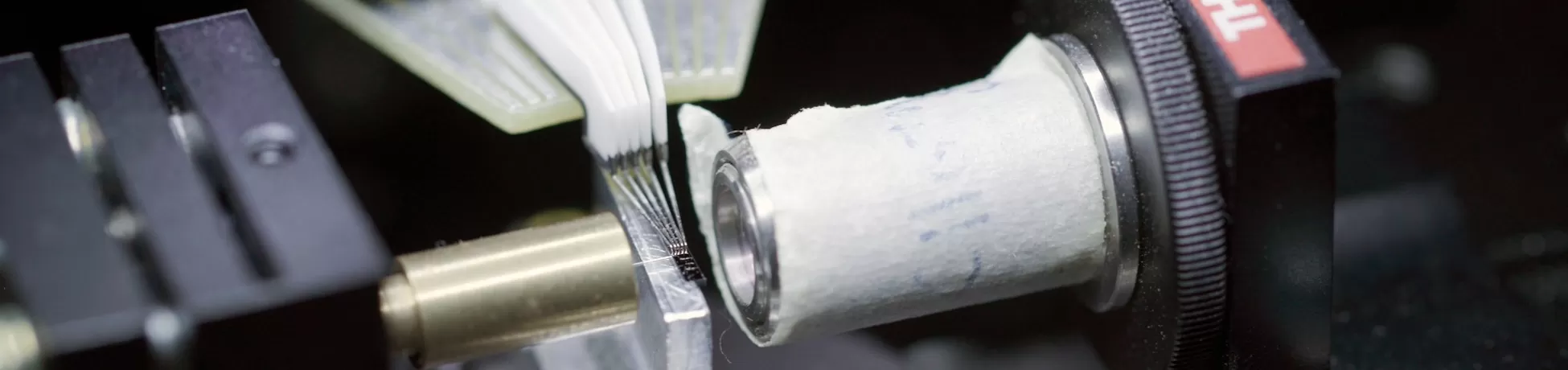 Close-up of a lab setup with a ThorLabs optical mount holding a small cylindrical component and a fan-shaped bundle of thin fiber-optic strands being aligned against a metal fixture.