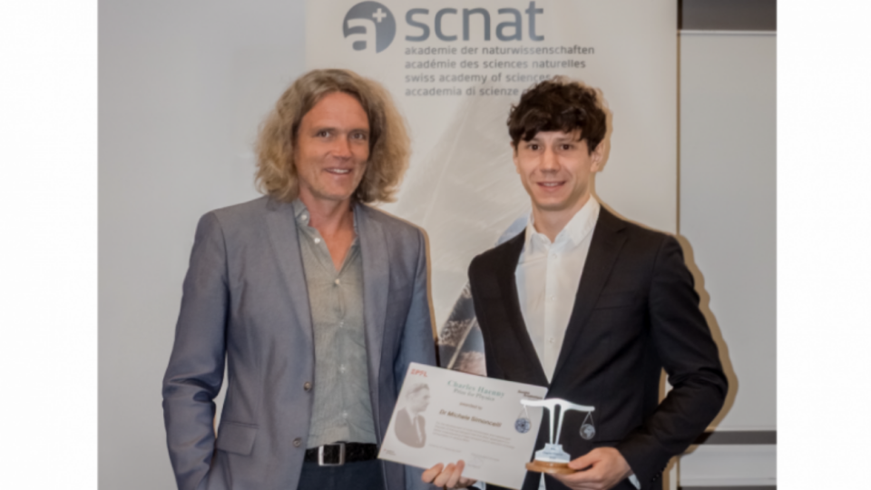 Two men stand together at an awards ceremony, with one holding a certificate and the other holding a trophy in front of a SCNAT backdrop.
