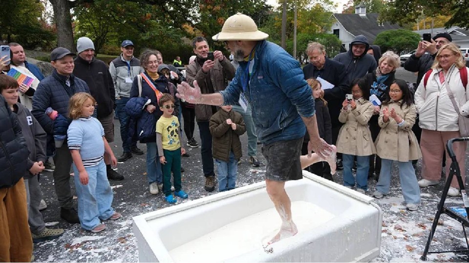 A man wearing shorts, a blue jacket, and a hat is dancing in a white bathtub of oobleck while adults and children look on from the sidelines.