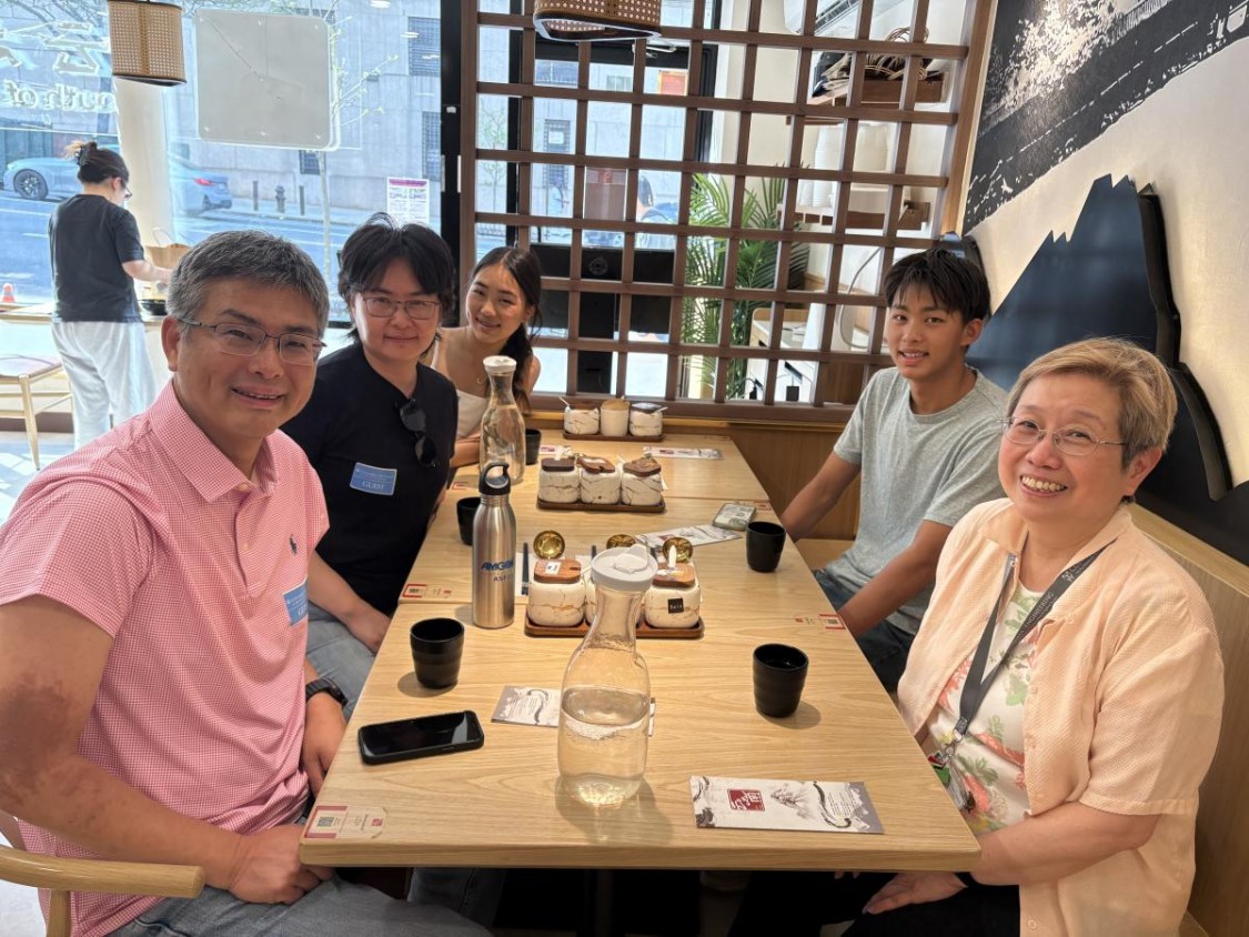Six people smiling around a wooden table in a modern café or restaurant space with brown lattice shelving and potted plants visible in the background, with water bottles, coffee cups, and printed materials on the table, and large windows overlooking a city street.